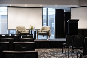 Un salón de conferencias con un escenario, dos sillones, una mesa con una planta y una pantalla de proyector.
