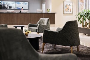 Un área de recepción moderna con un mostrador de madera, dos sillones grises, una mesa redonda de centro y una planta en maceta.