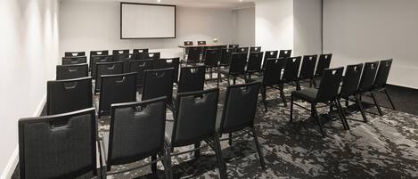 A conference room with rows of black chairs, a large table, a projector screen, and a carpeted floor.