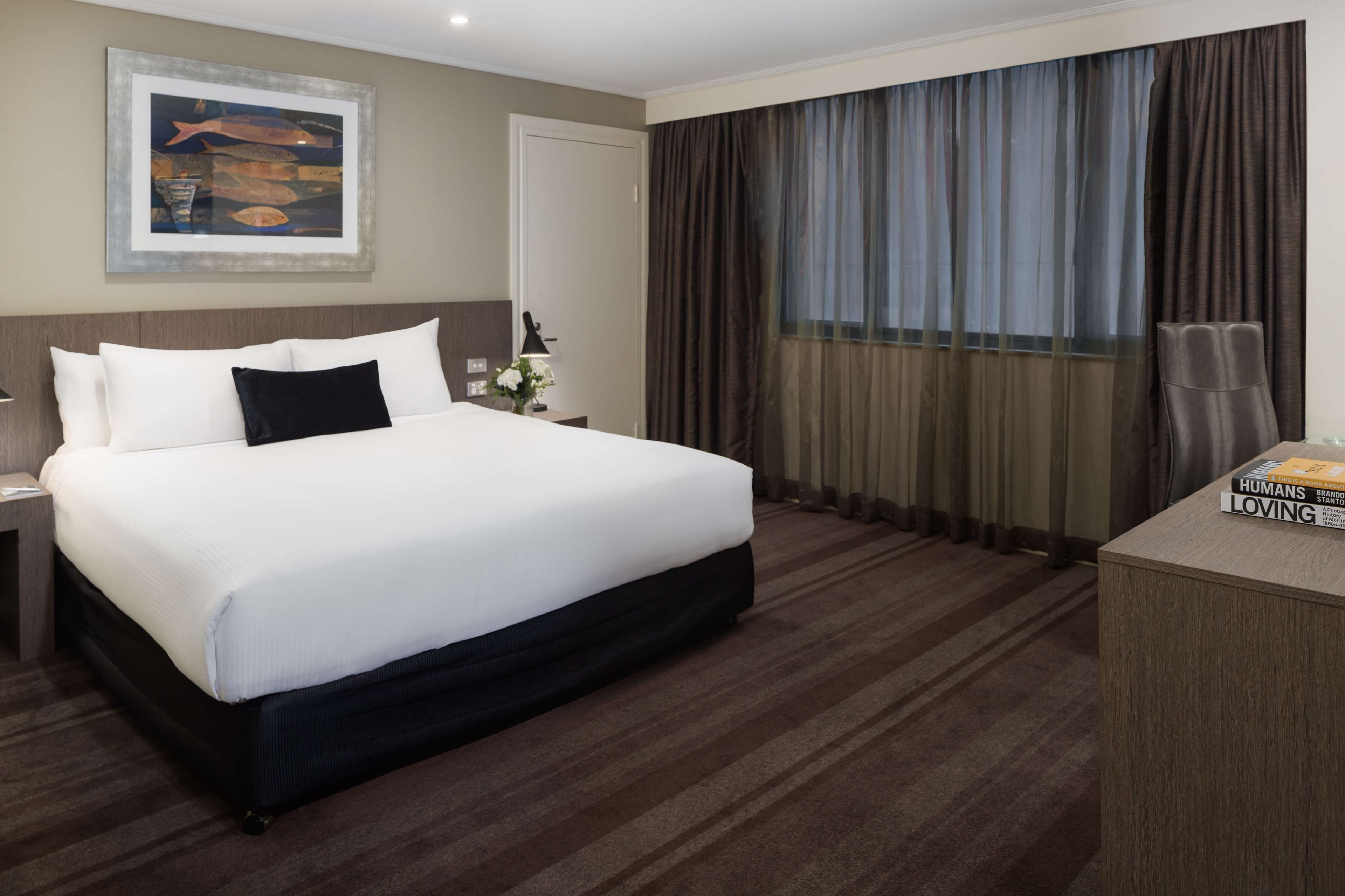 A hotel room with a large bed, a nightstand, a desk with a lamp, a chair, and a window with curtains.