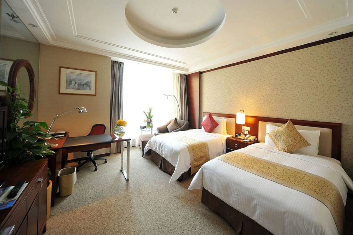 Quarto de hotel com duas camas, uma escrivaninha, uma cadeira, um abajur, um telefone e uma planta.