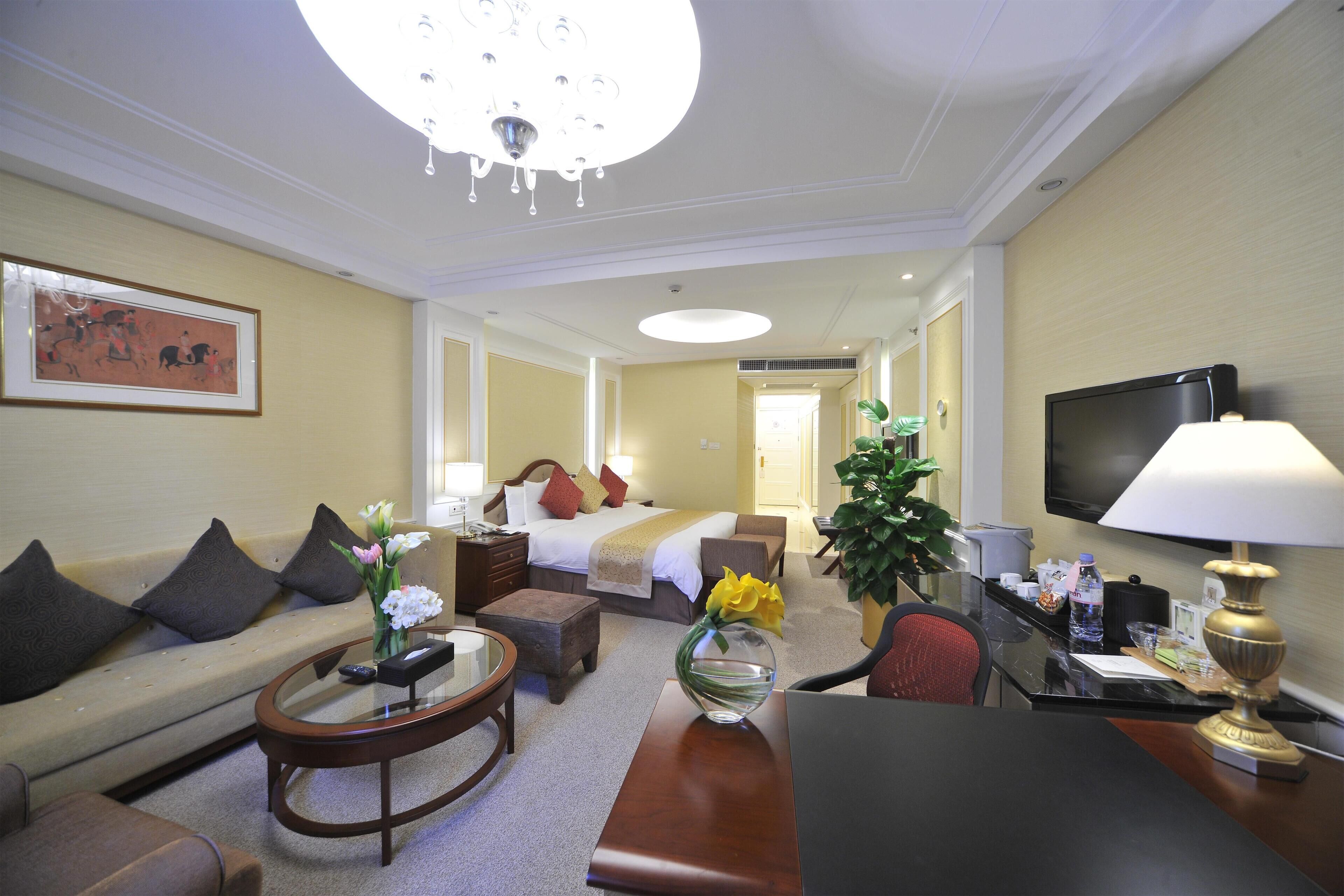 A hotel room with a sofa, a round coffee table, a bed with pillows, a TV, a desk with a lamp, and a plant.