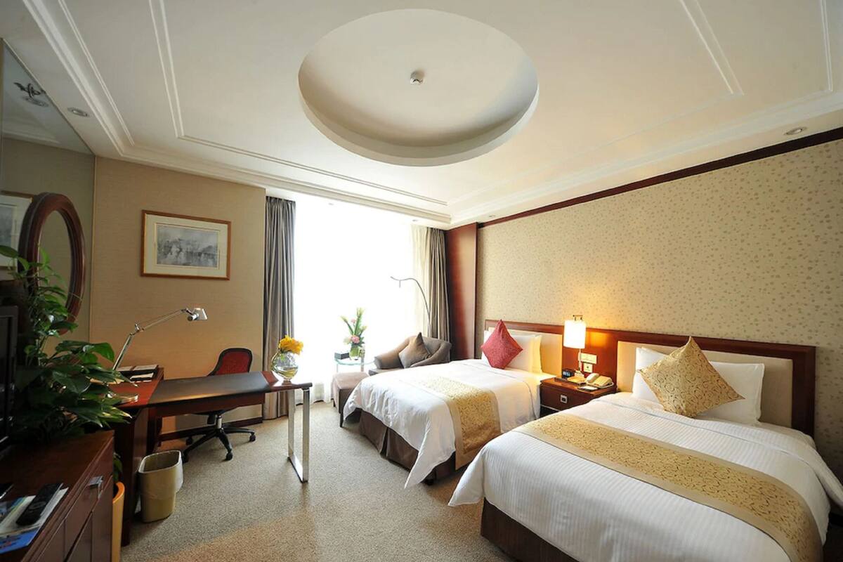Quarto de hotel com duas camas, uma escrivaninha, uma cadeira, um abajur, um espelho e uma planta.