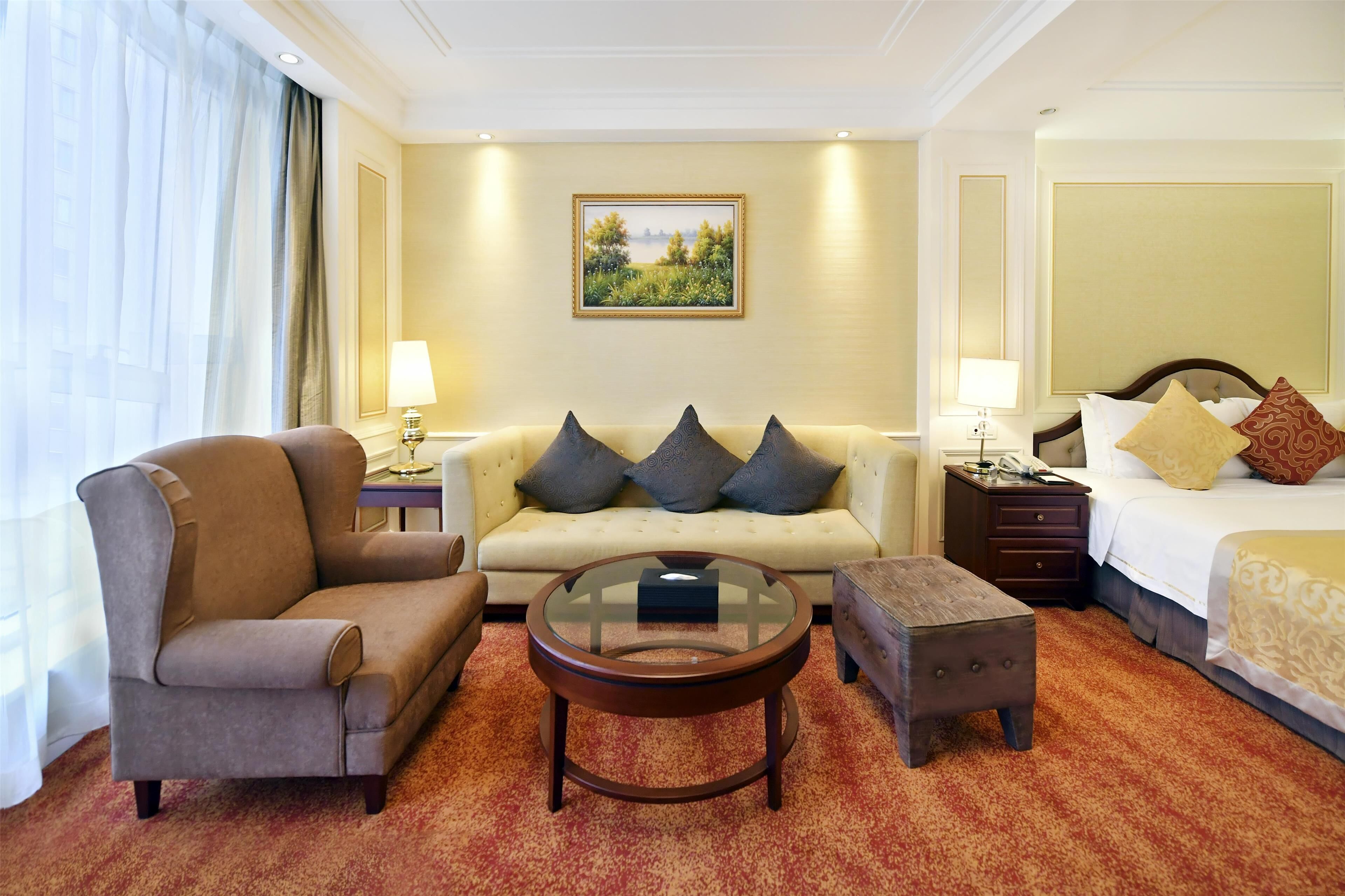 Een hotelkamer met een bank, fauteuil, rond bijzettafeltje, poef en een bed met kussens.