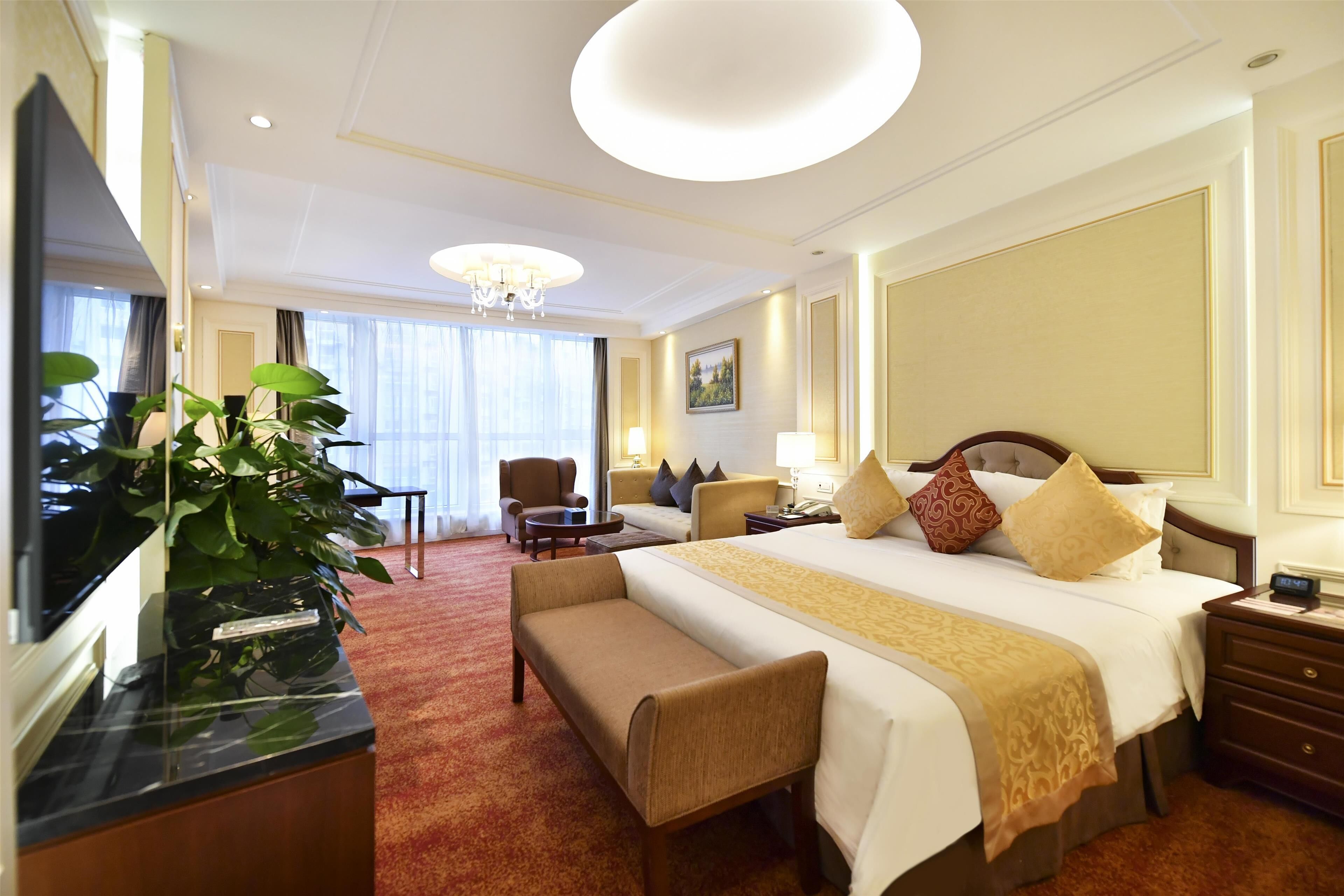 Une chambre d’hôtel avec un grand lit, un bureau, une chaise, un banc, une télévision et une grande fenêtre.
