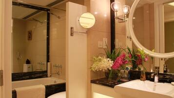 A modern bathroom with a bathtub, toilet, and sink. There are two mirrors, one round and one oval. The walls are tiled, and there is a vase with flowers on the sink counter.