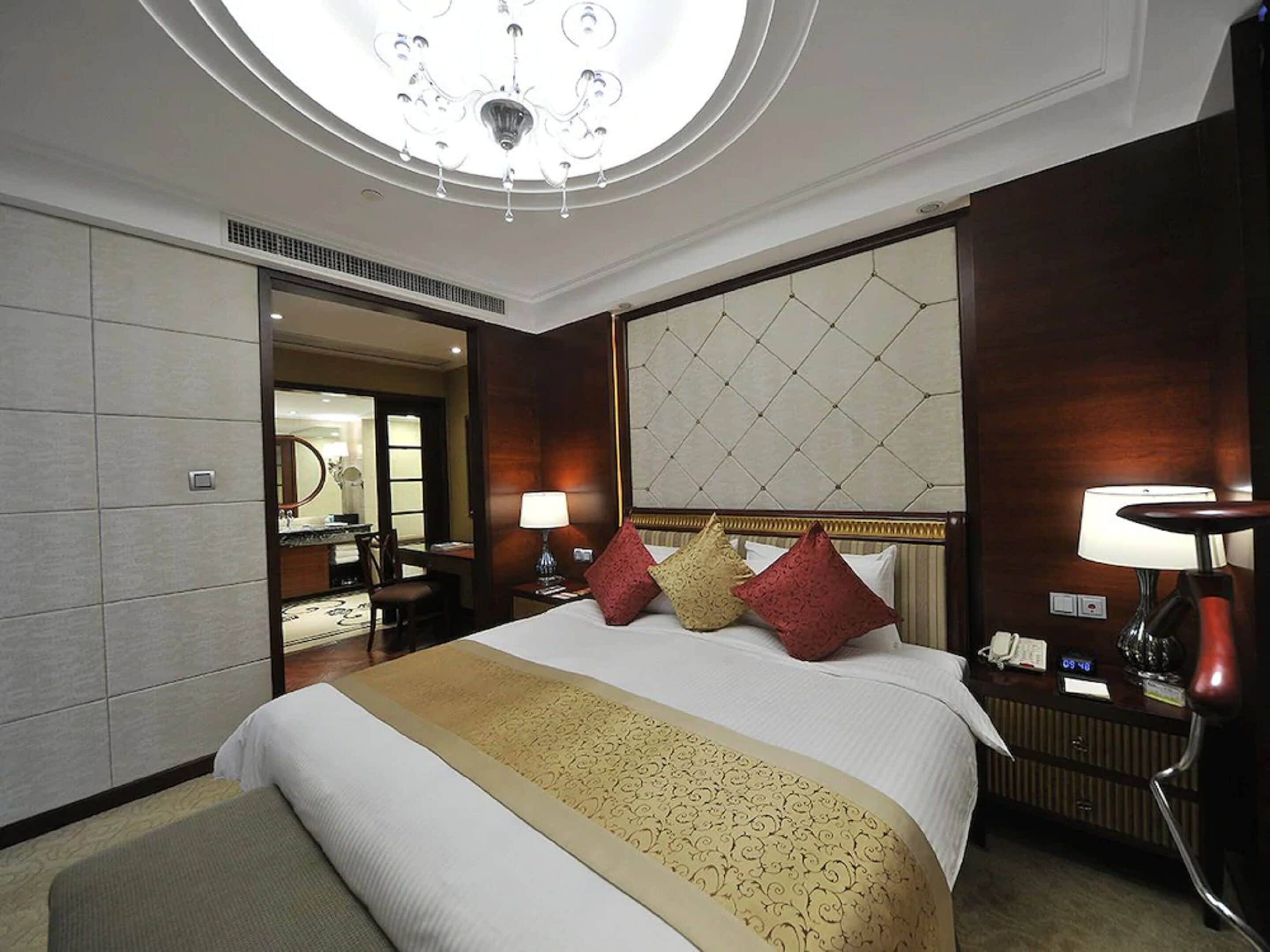 A hotel room with a large bed, a chandelier, a mirror, and a bedside table with a lamp.