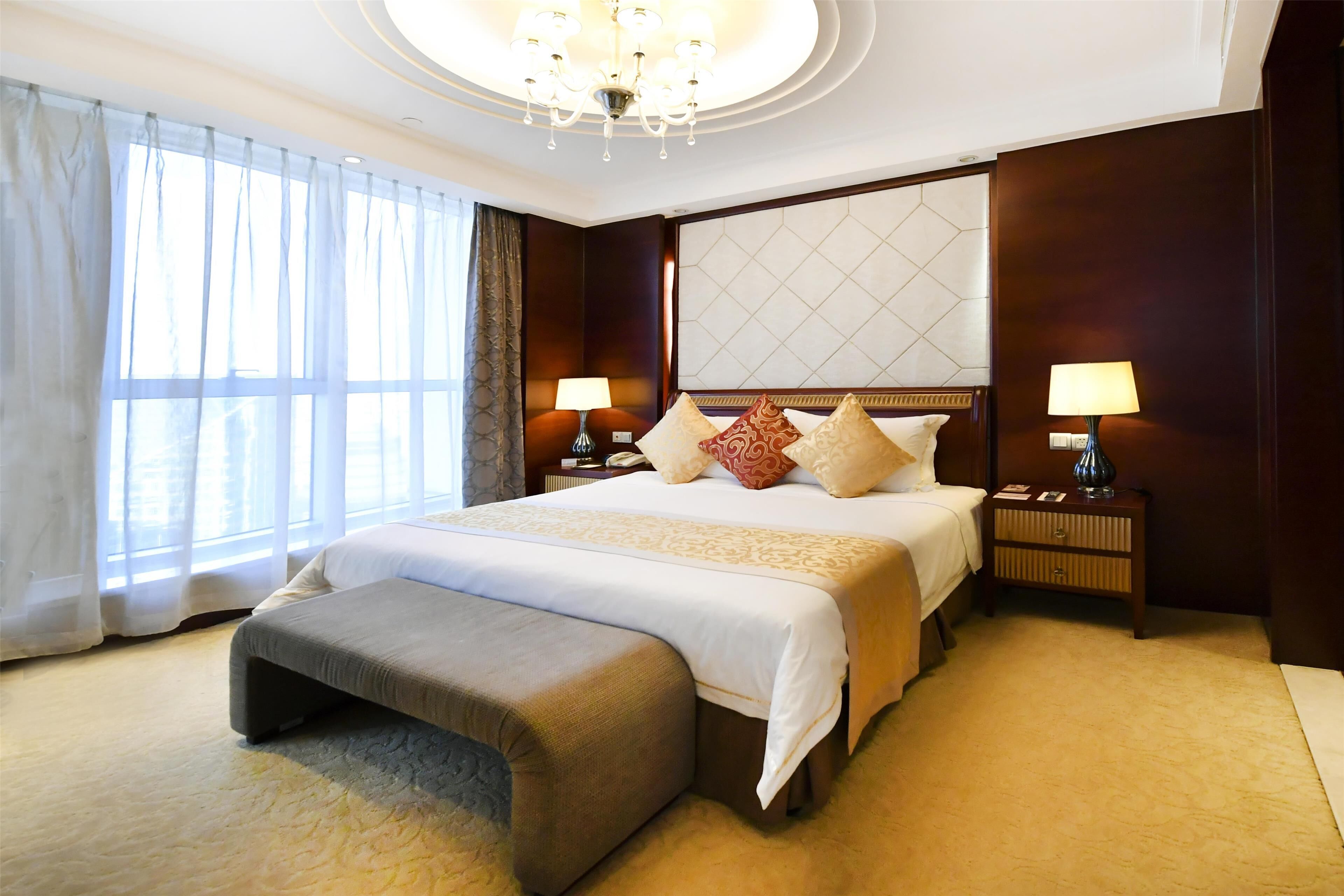 A hotel room with a large bed, two bedside tables with lamps, a bench, and a large window with curtains.