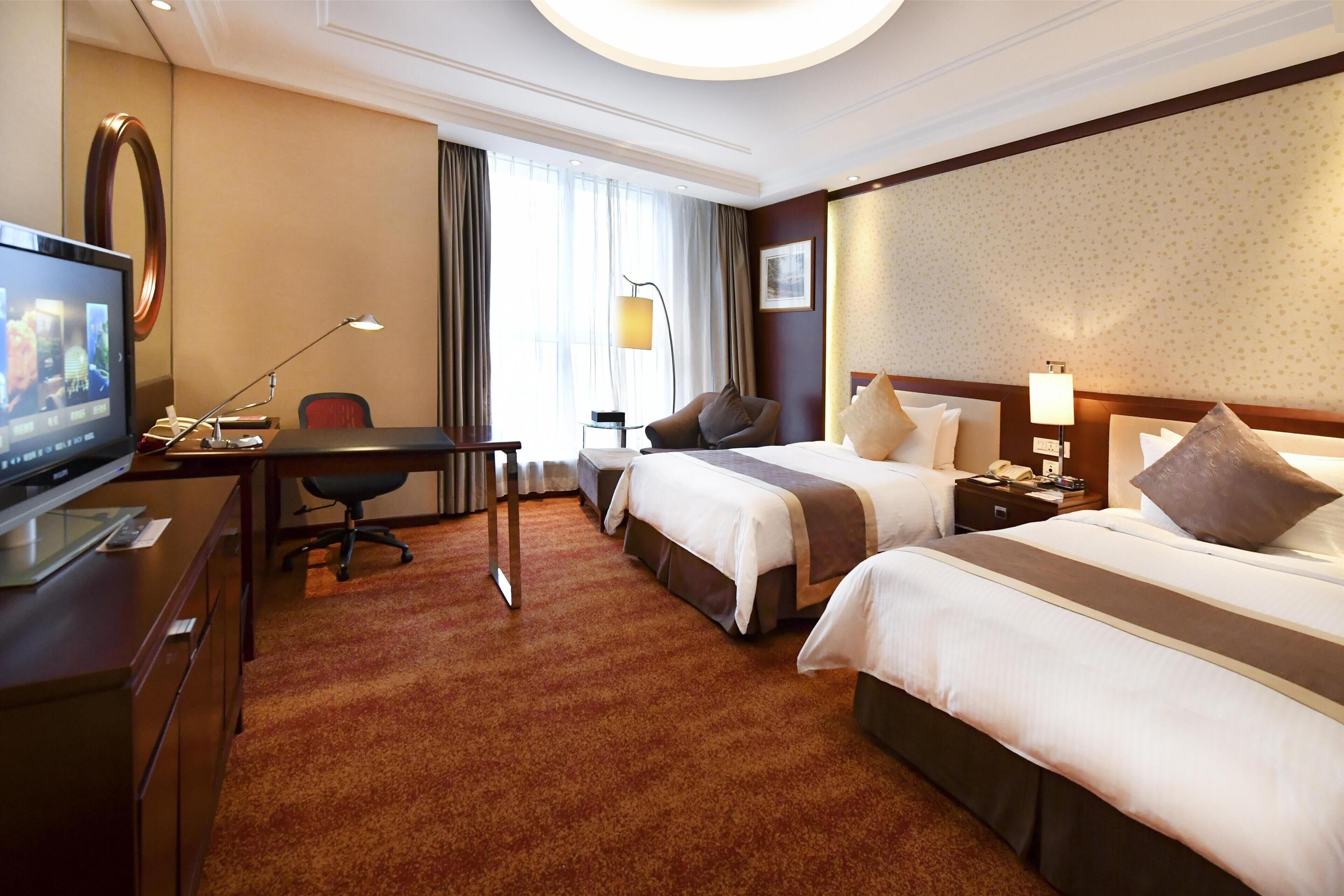 Ein Hotelzimmer mit zwei Betten, einem Schreibtisch mit Computer, einem Fernseher und einem großen Fenster mit Vorhängen.