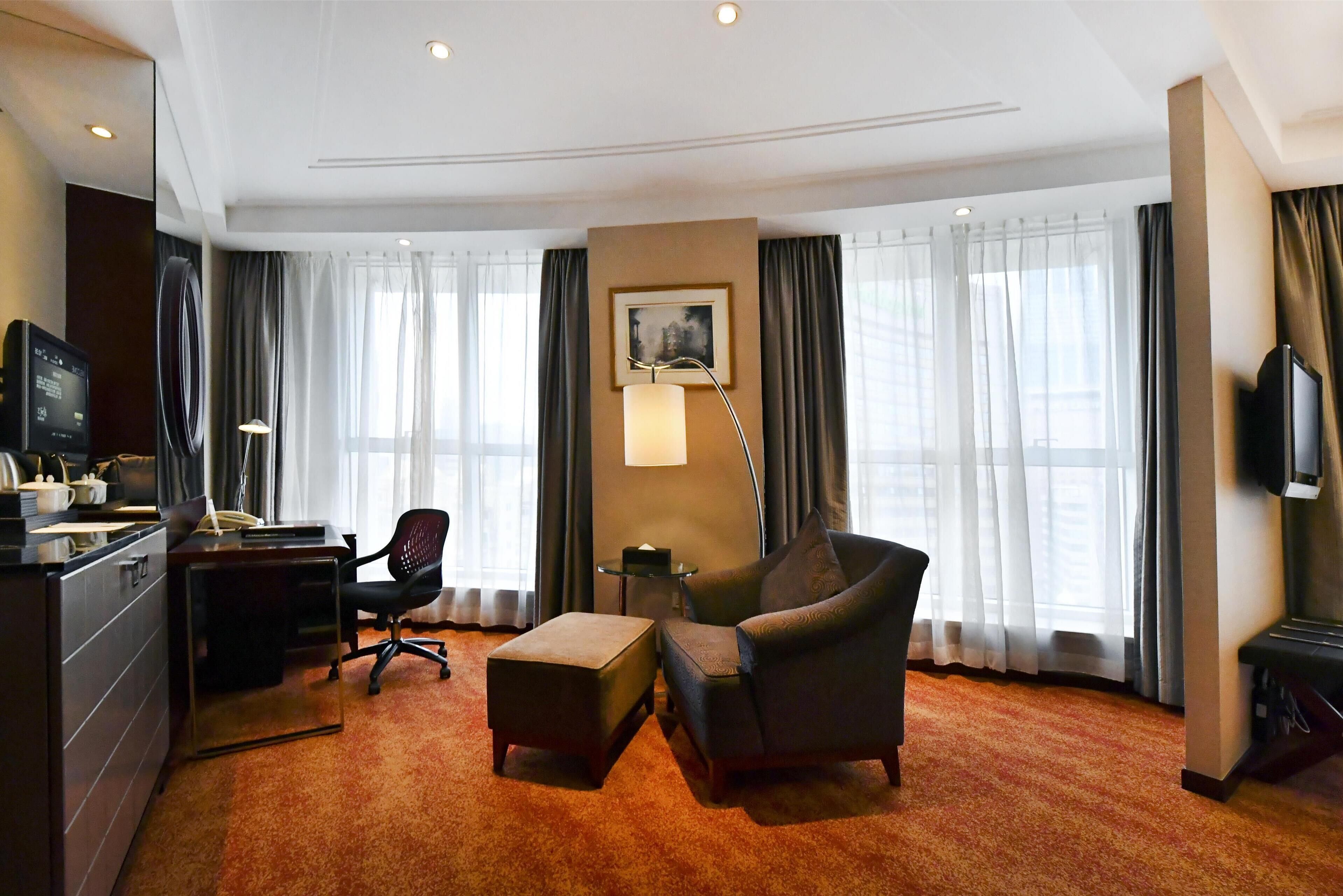 Hotelkamer met een bureau, stoel, poef, grote ramen met gordijnen, een flatscreen televisie en een lamp.
