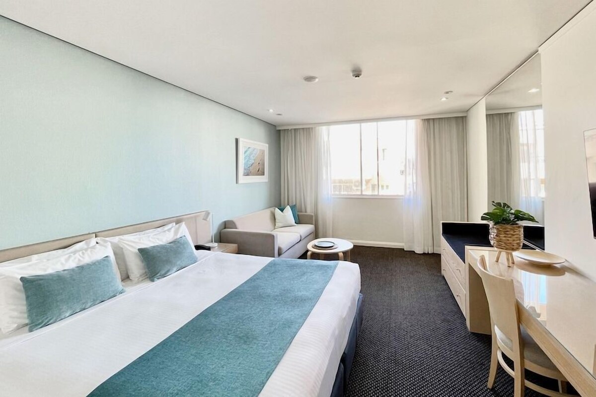 A hotel room with a bed, a desk, a chair, a small table, and a TV.