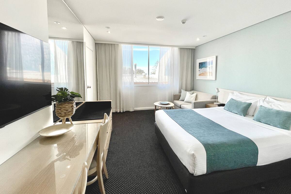 A modern hotel room with a large bed, a desk with a laptop, a flat-screen TV, and a view of the city.