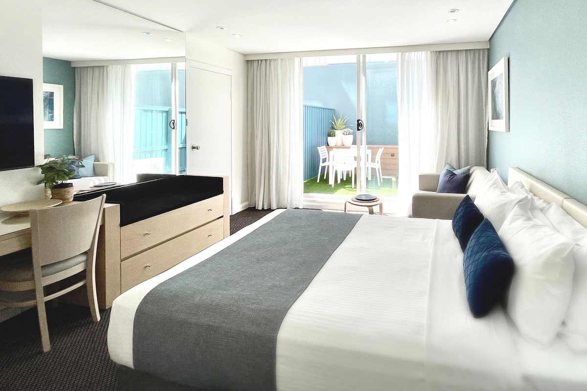 A modern hotel room with a large bed, a desk, and a balcony with a table and chairs.