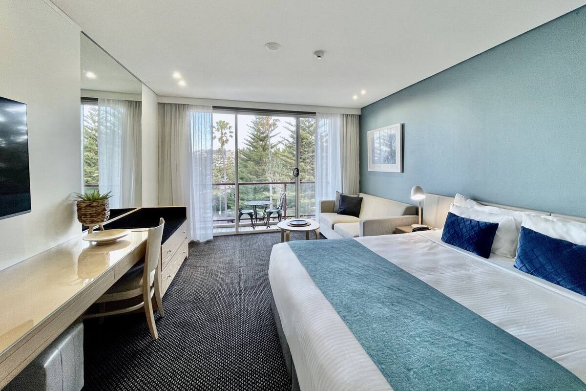 A modern hotel room with a large bed, a desk, a chair, a small table, and a balcony with a view of trees.