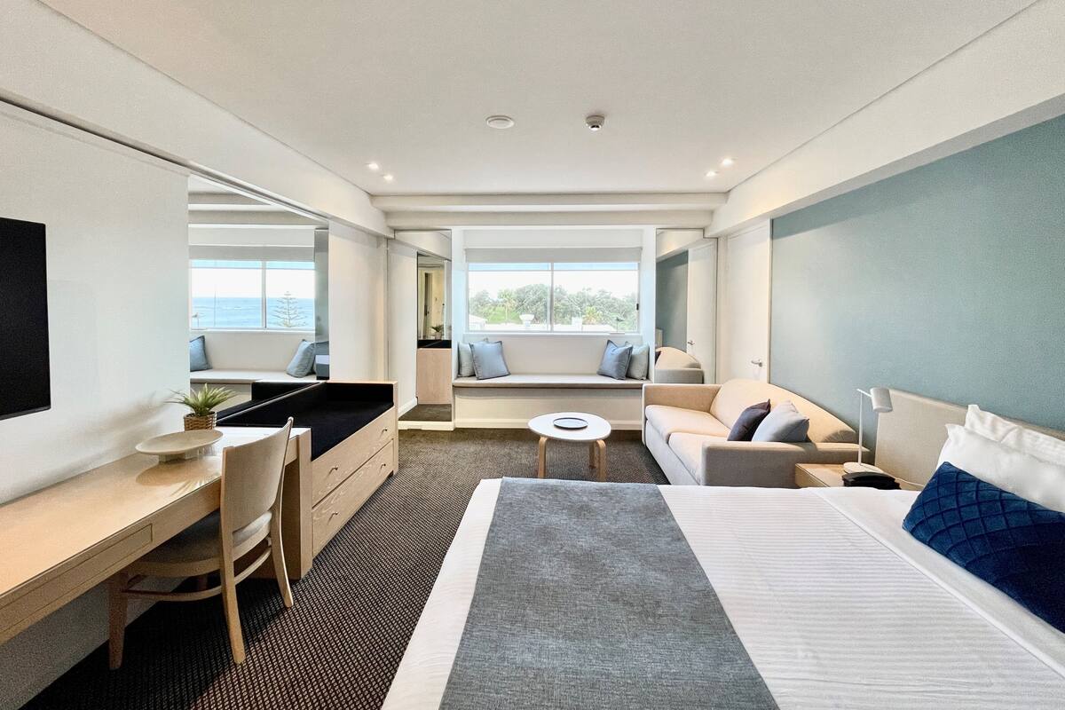 A modern hotel room with a large bed, a desk, a sofa, and a view of the sea.