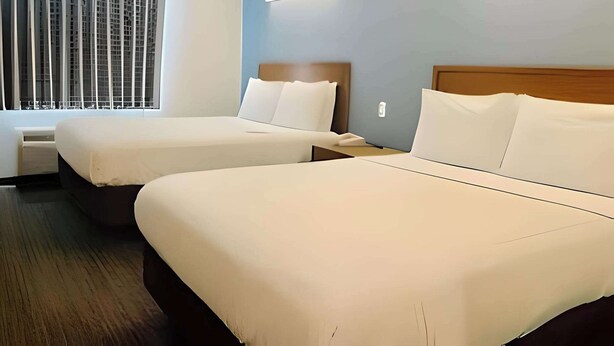 A hotel room with two beds, a window with blinds, a bedside table, and a wall-mounted light fixture.