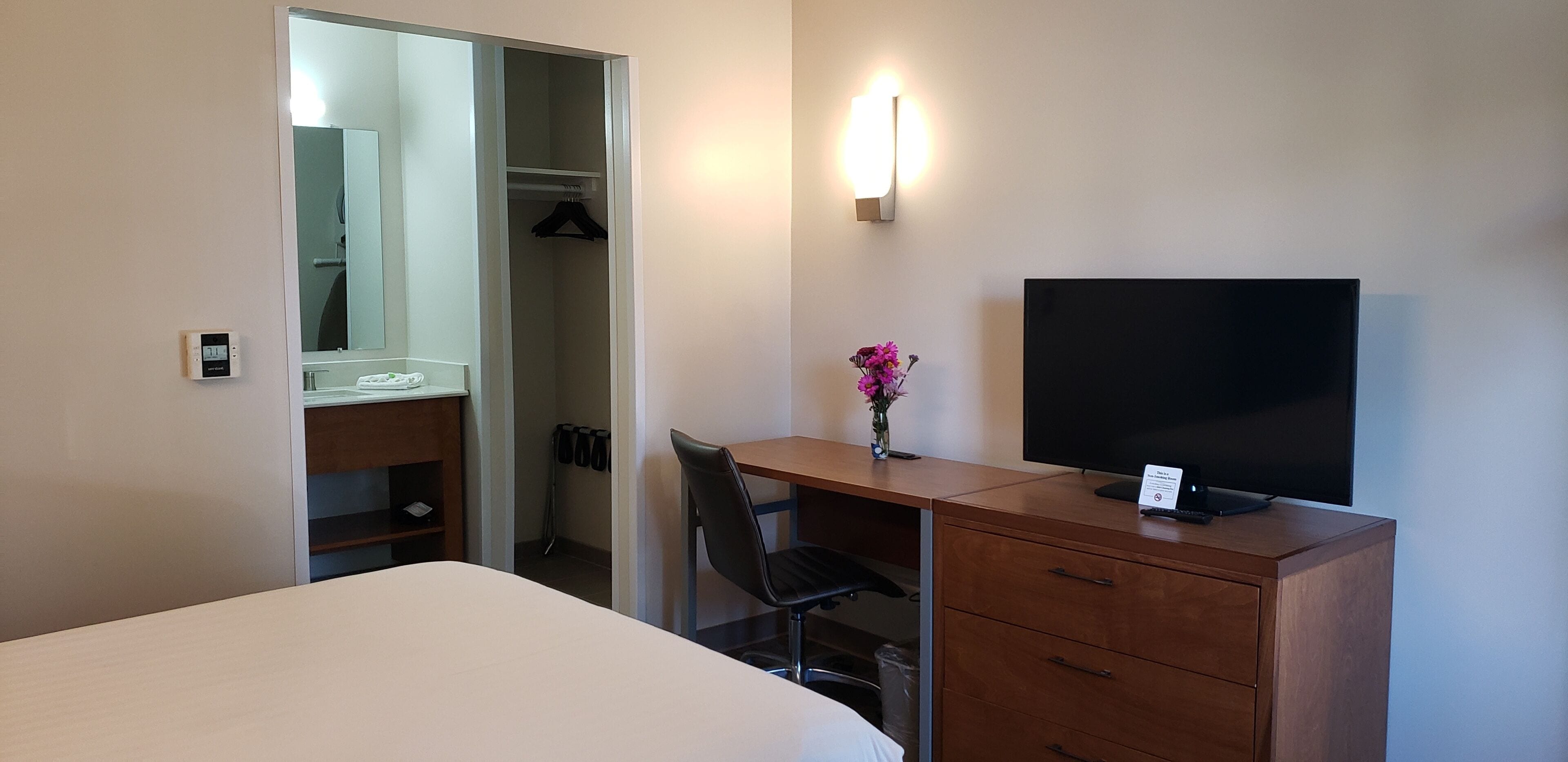 Habitación de hotel con cama, escritorio con silla, televisor, espejo, armario y lámpara fijada a la pared.