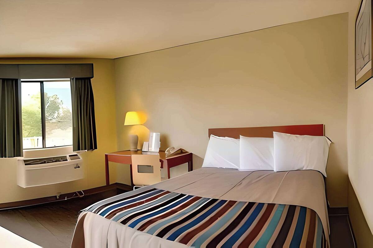 A hotel room with a bed, a desk, a chair, a lamp, and a window with curtains.