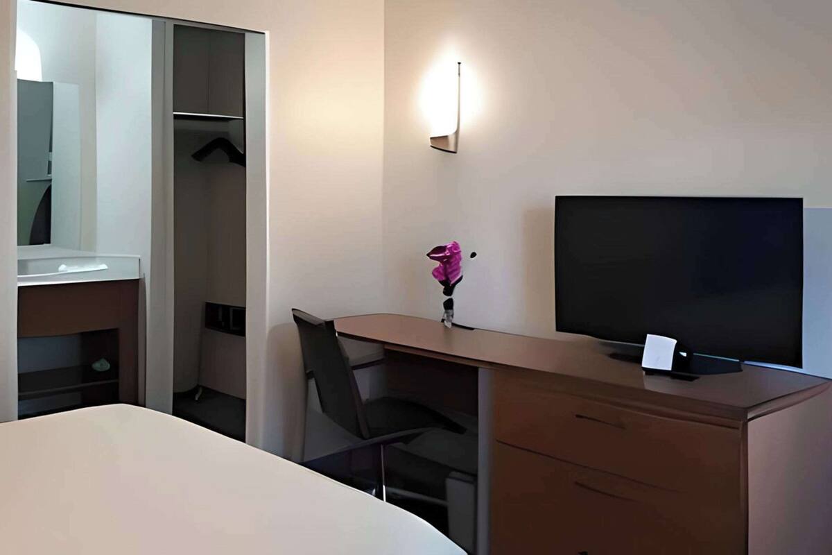 A hotel room with a bed, a desk with a chair, a TV, a wardrobe, and a wall-mounted light.