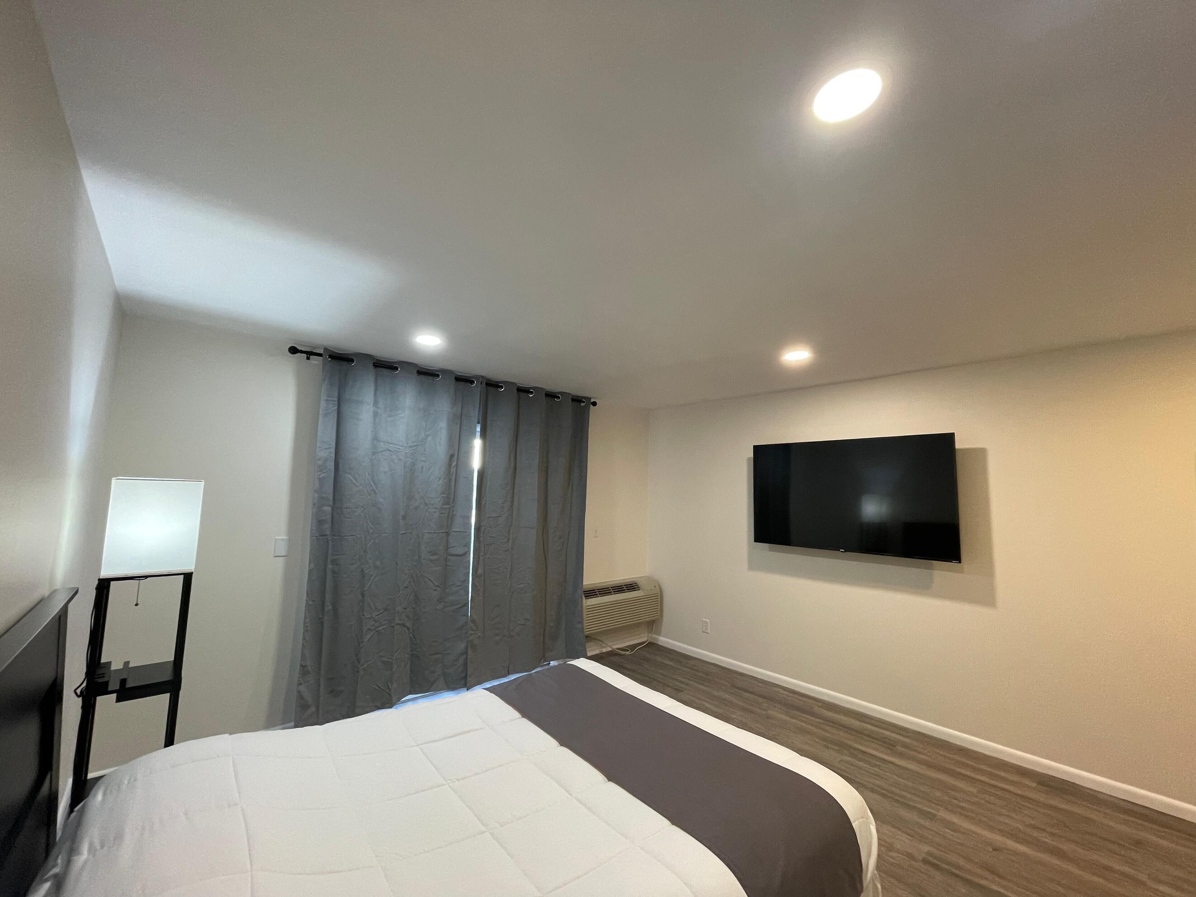 A hotel room with a bed, a flat-screen TV mounted on the wall, a ceiling light, and a window with curtains.