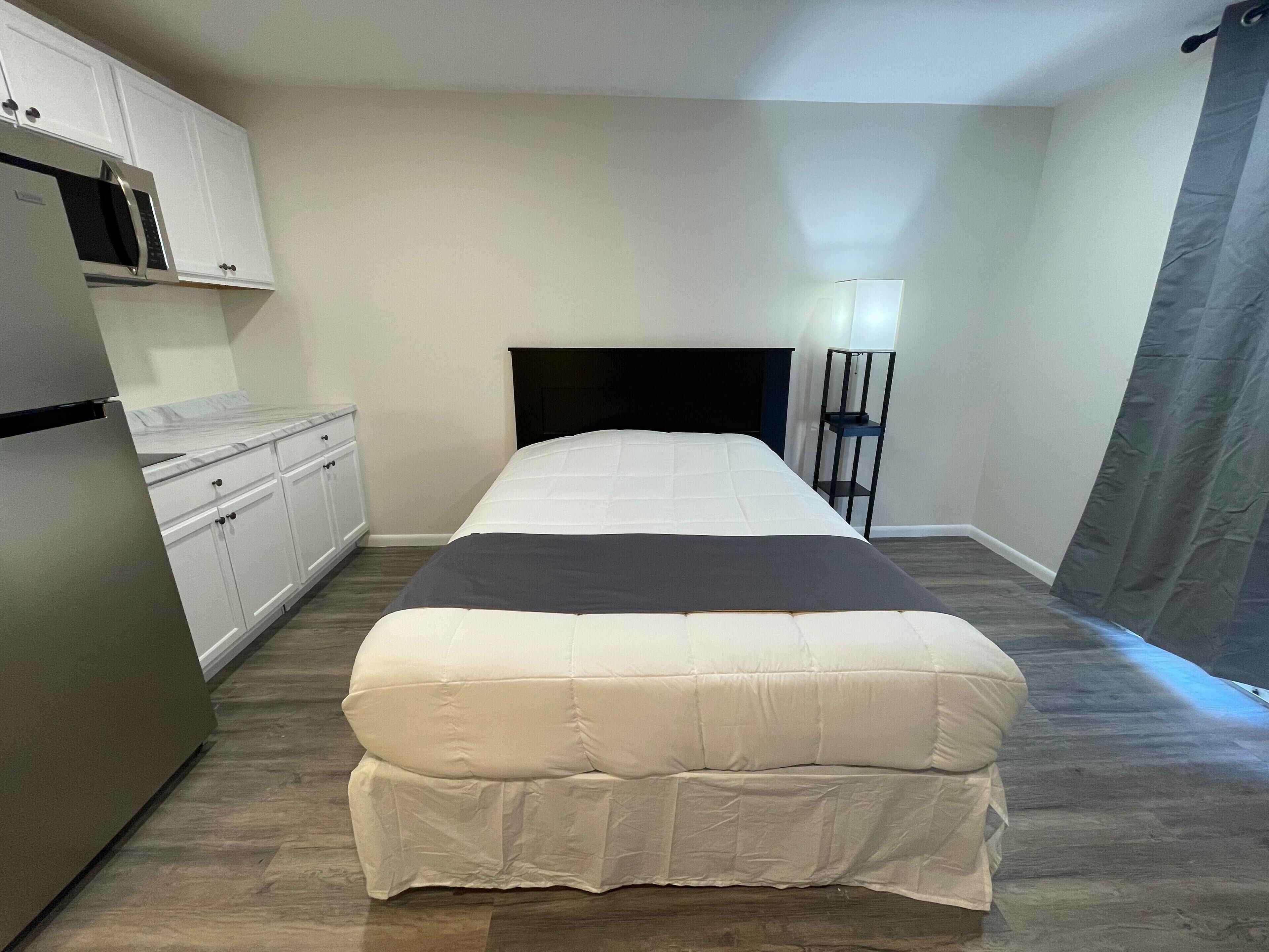 A bedroom with a bed, a nightstand, a lamp, and a kitchenette.