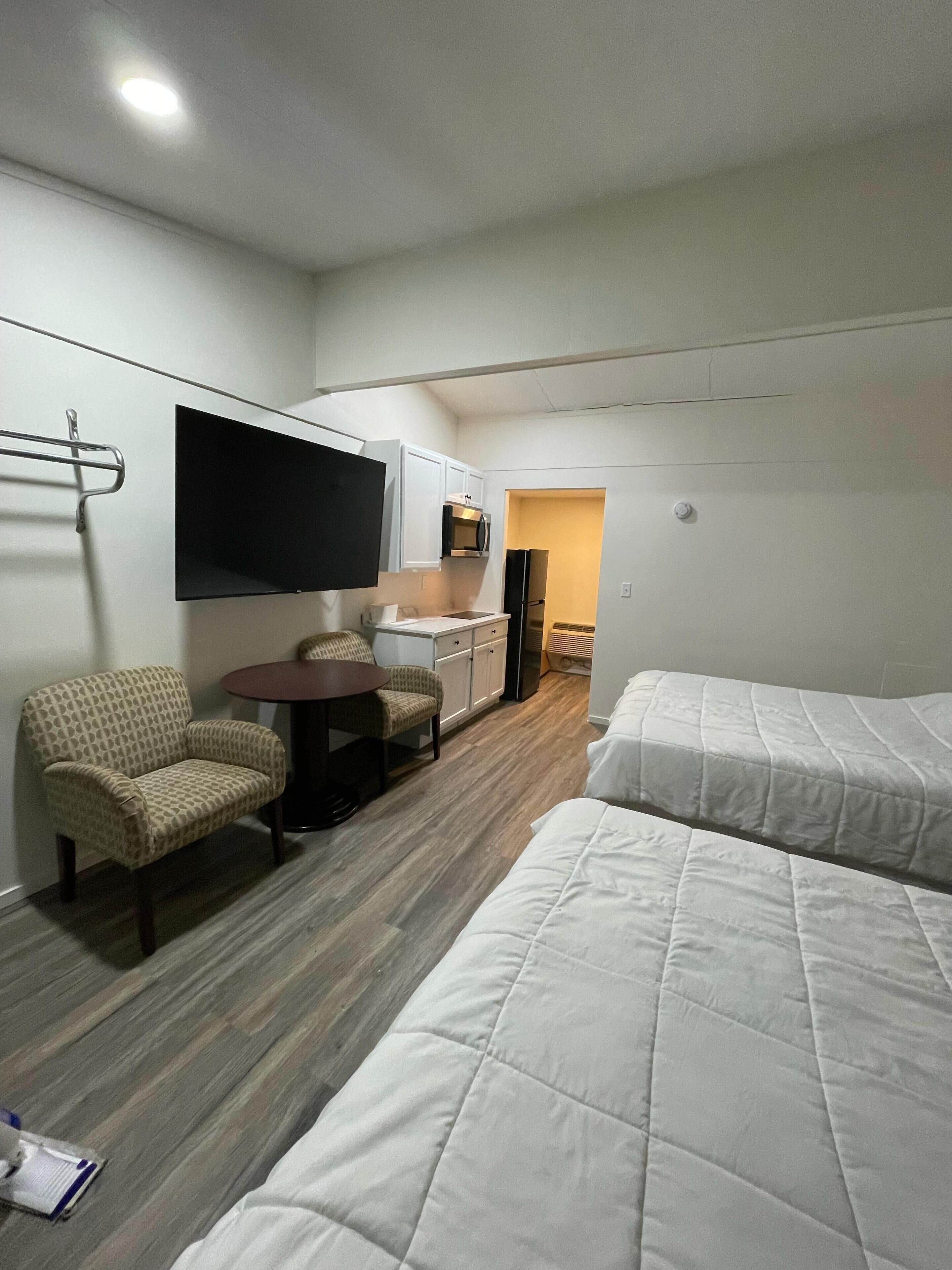 A hotel room with two beds, a TV mounted on the wall, a kitchenette, and a seating area with a chair and table.