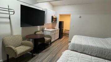 A hotel room with two beds, a TV mounted on the wall, a kitchenette, and a seating area with a chair and table.