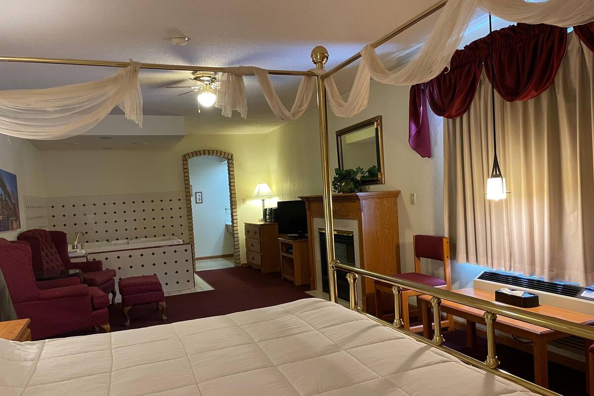 A hotel room with a four-poster bed, a sitting area with a sofa and armchairs, a television, a fireplace, and a large window with curtains.