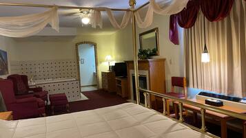 Quarto de hotel com cama de dossel, área de estar com sofá e poltronas, televisão, lareira e uma grande janela com cortinas.