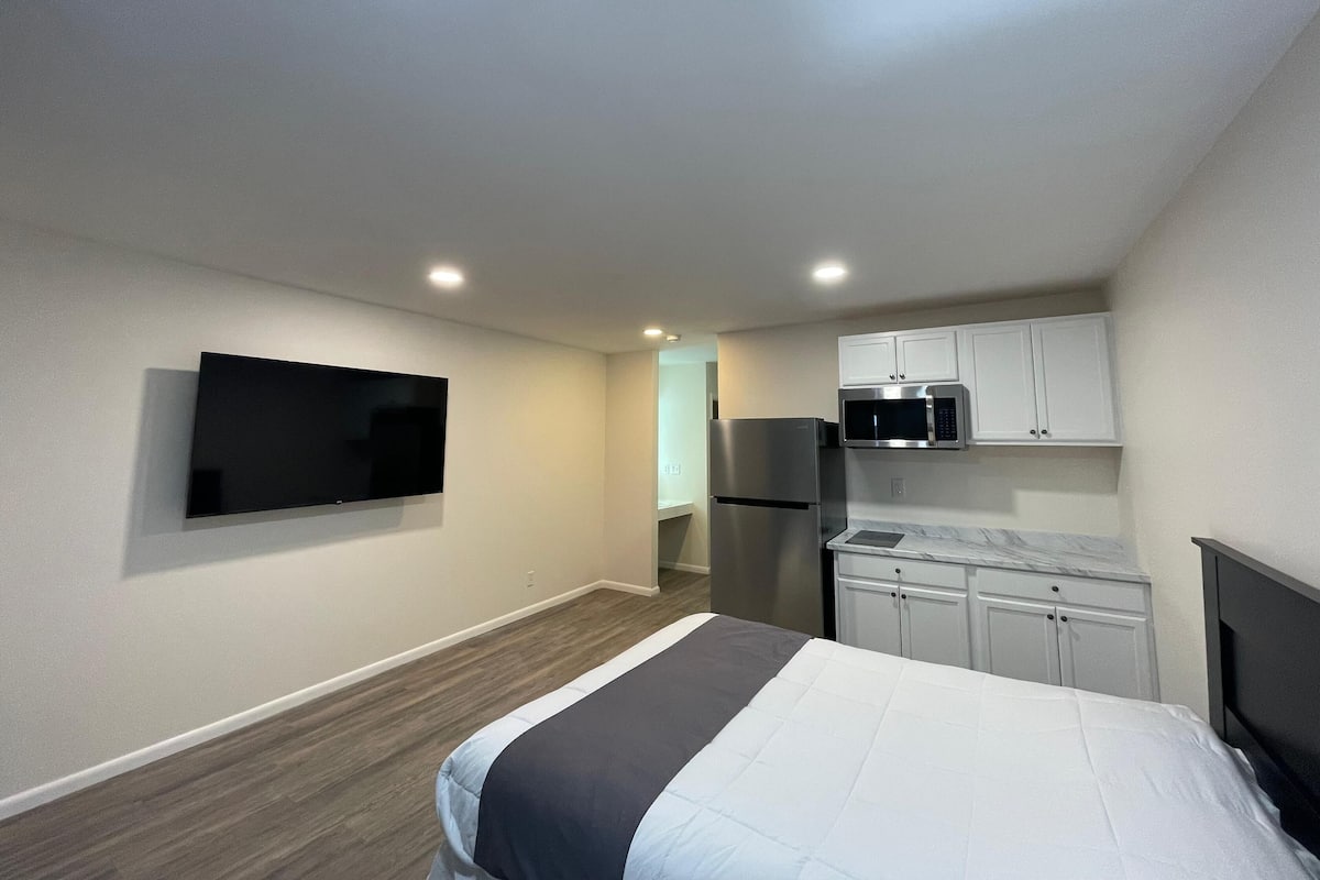 A compact hotel room with a bed, a flat-screen TV, a kitchenette with a fridge and microwave, and a small bathroom.