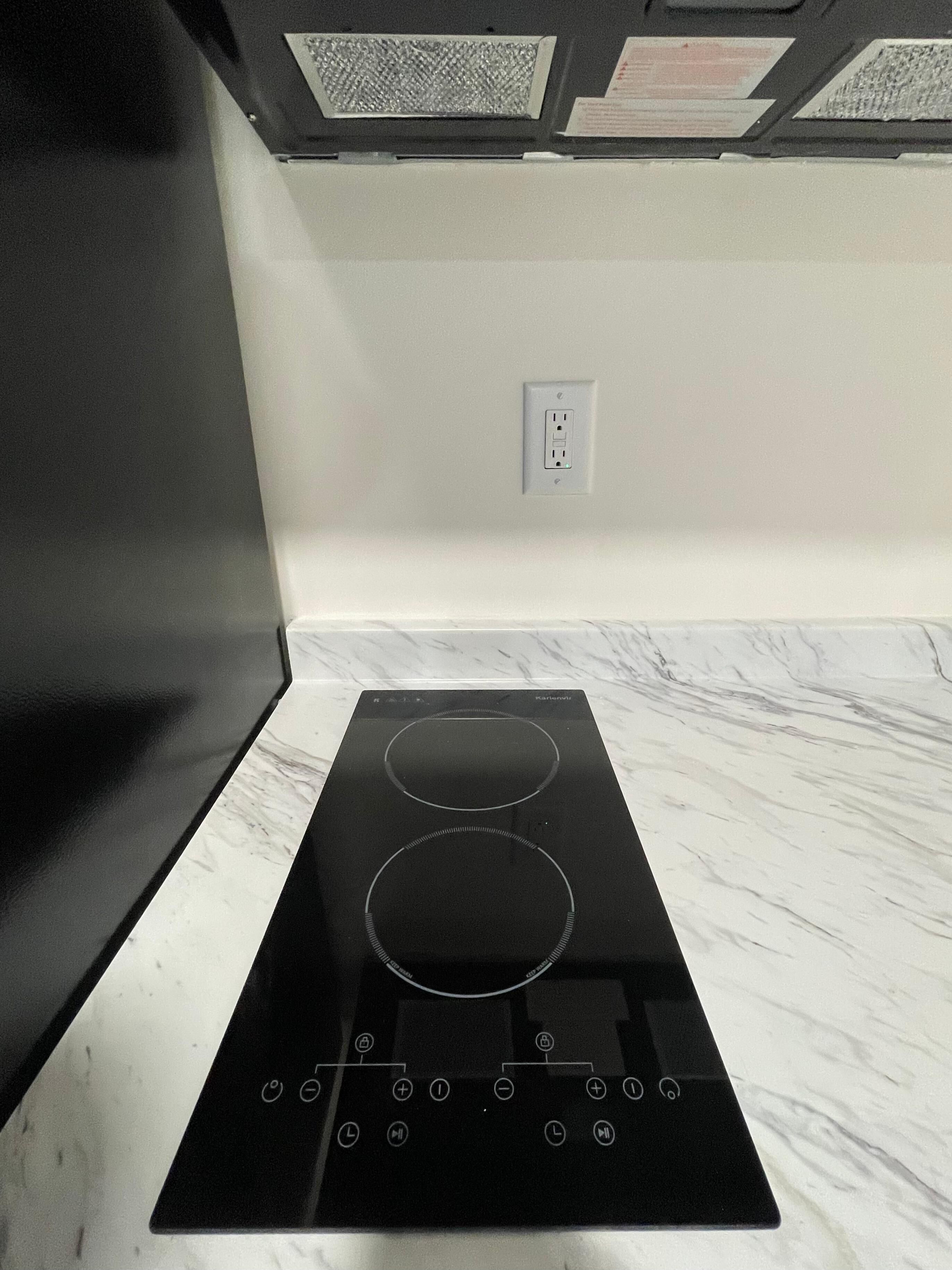 A black induction cooktop with two burners on a white marble countertop, a power outlet on the wall, and a vent above the cooktop.