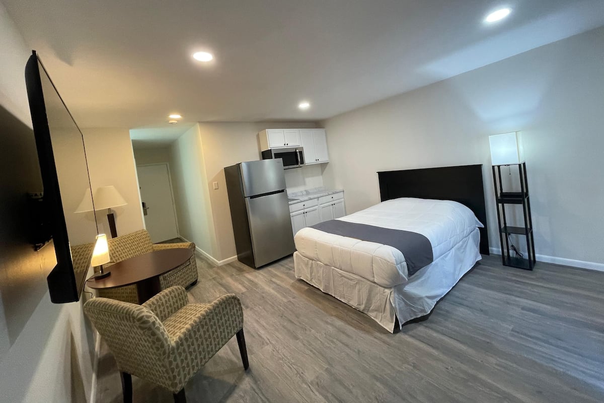A hotel room with a bed, a desk with a lamp, a refrigerator, a microwave, a chair, and a floor lamp.