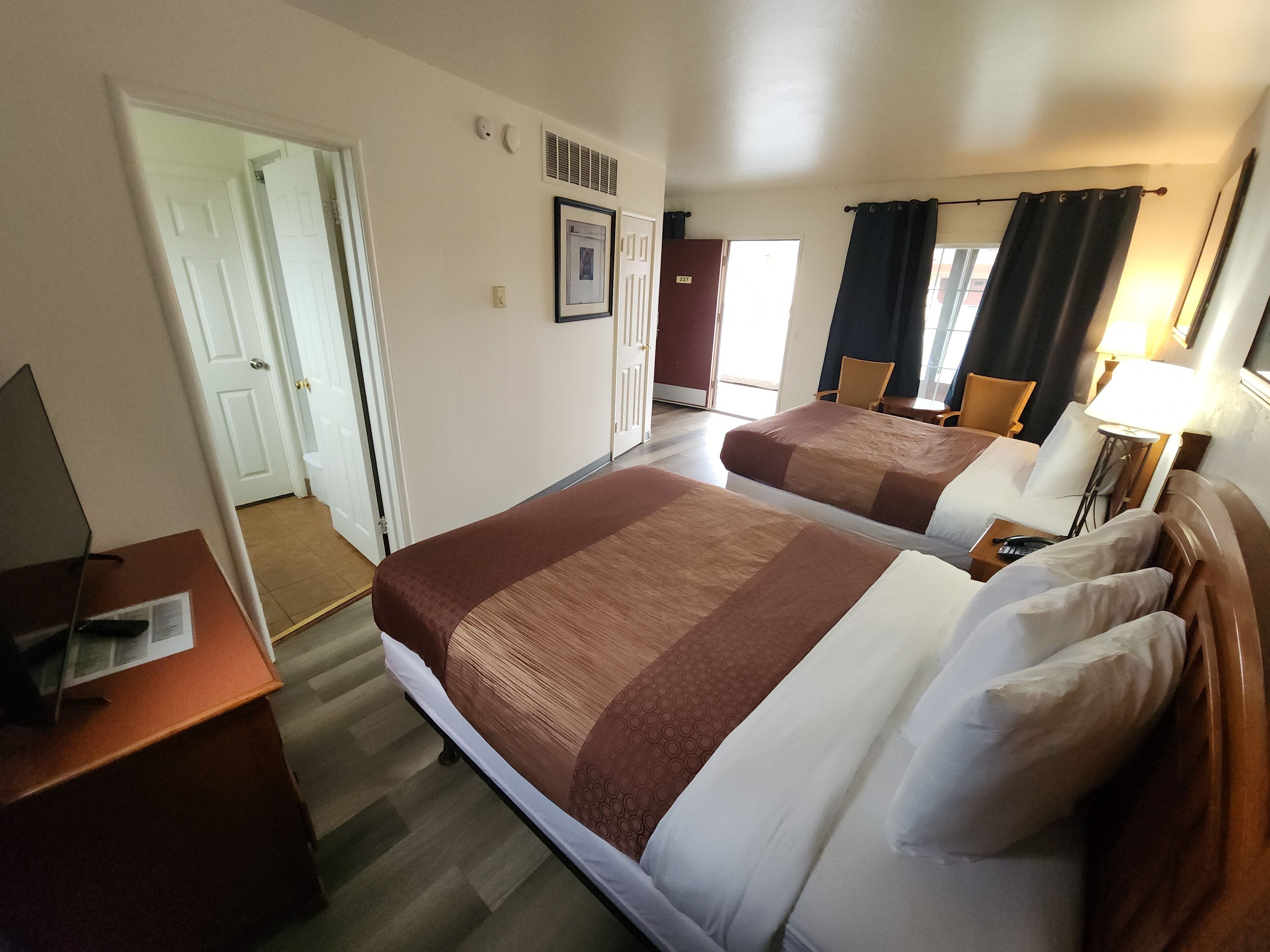 A hotel room with two beds, a desk, a chair, a lamp, and a door leading to another room.