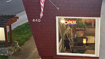 Un bâtiment en briques surmonté d’un drapeau américain, une devanture de magasin avec une enseigne indiquant « OUVERT », et une fenêtre où l’on aperçoit une plante et une lampe.