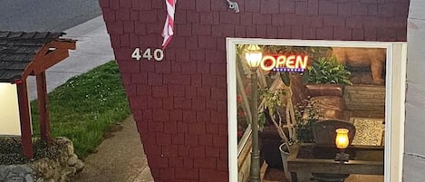 Un bâtiment en briques surmonté d’un drapeau américain, une devanture de magasin avec une enseigne indiquant « OUVERT », et une fenêtre où l’on aperçoit une plante et une lampe.