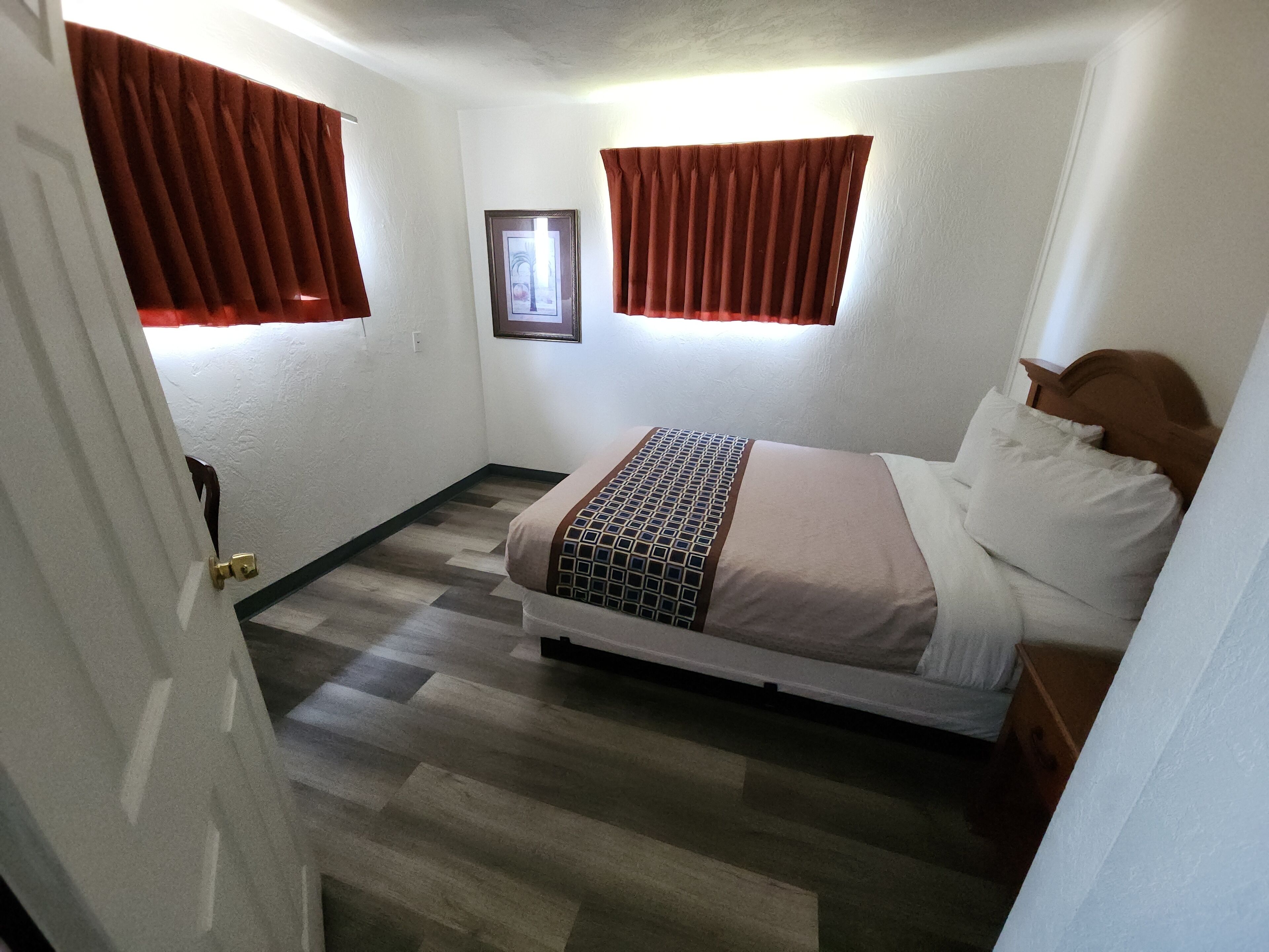 Een slaapkamer met een bed, een houten hoofdbord, een ingelijste foto aan de muur en een raam met gordijnen.