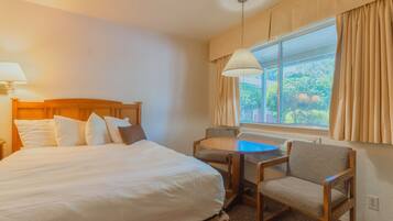 A hotel room with a bed, a table with chairs, a window with curtains, and a lamp.