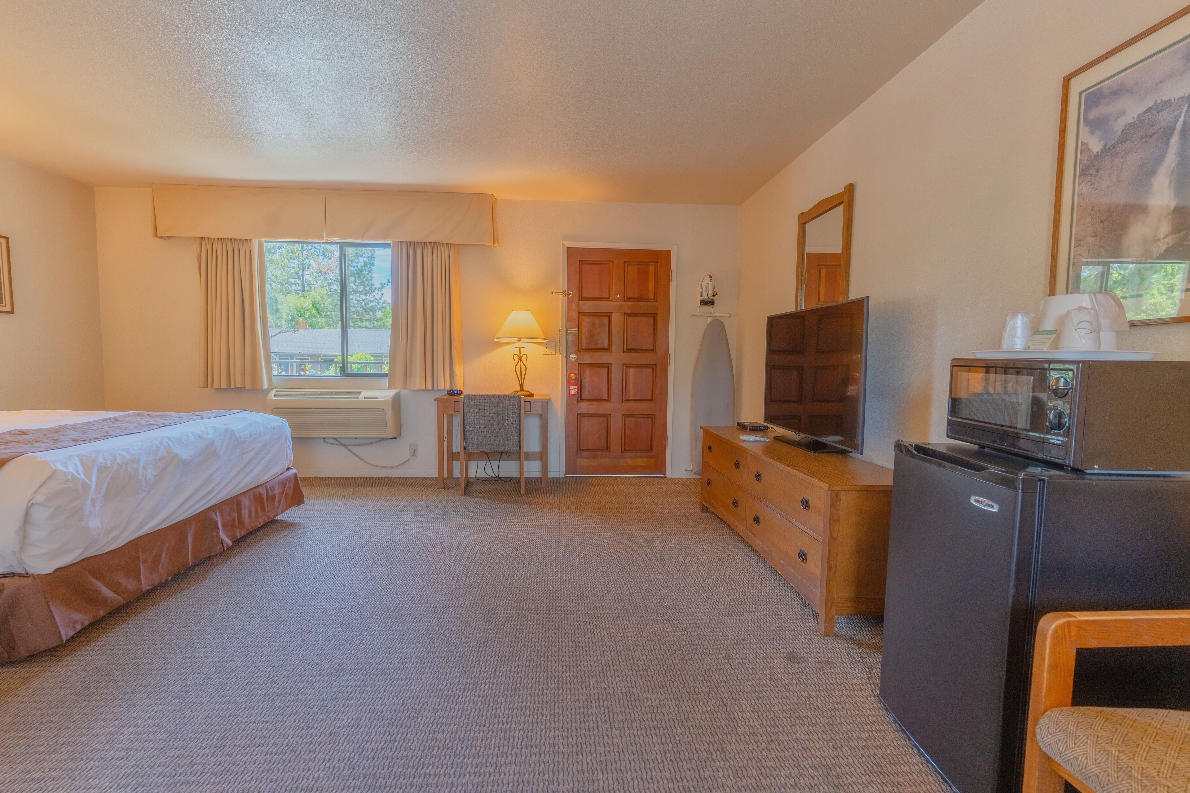 A hotel room with a bed, a desk, a chair, a dresser, a television, a refrigerator, and a window with curtains.