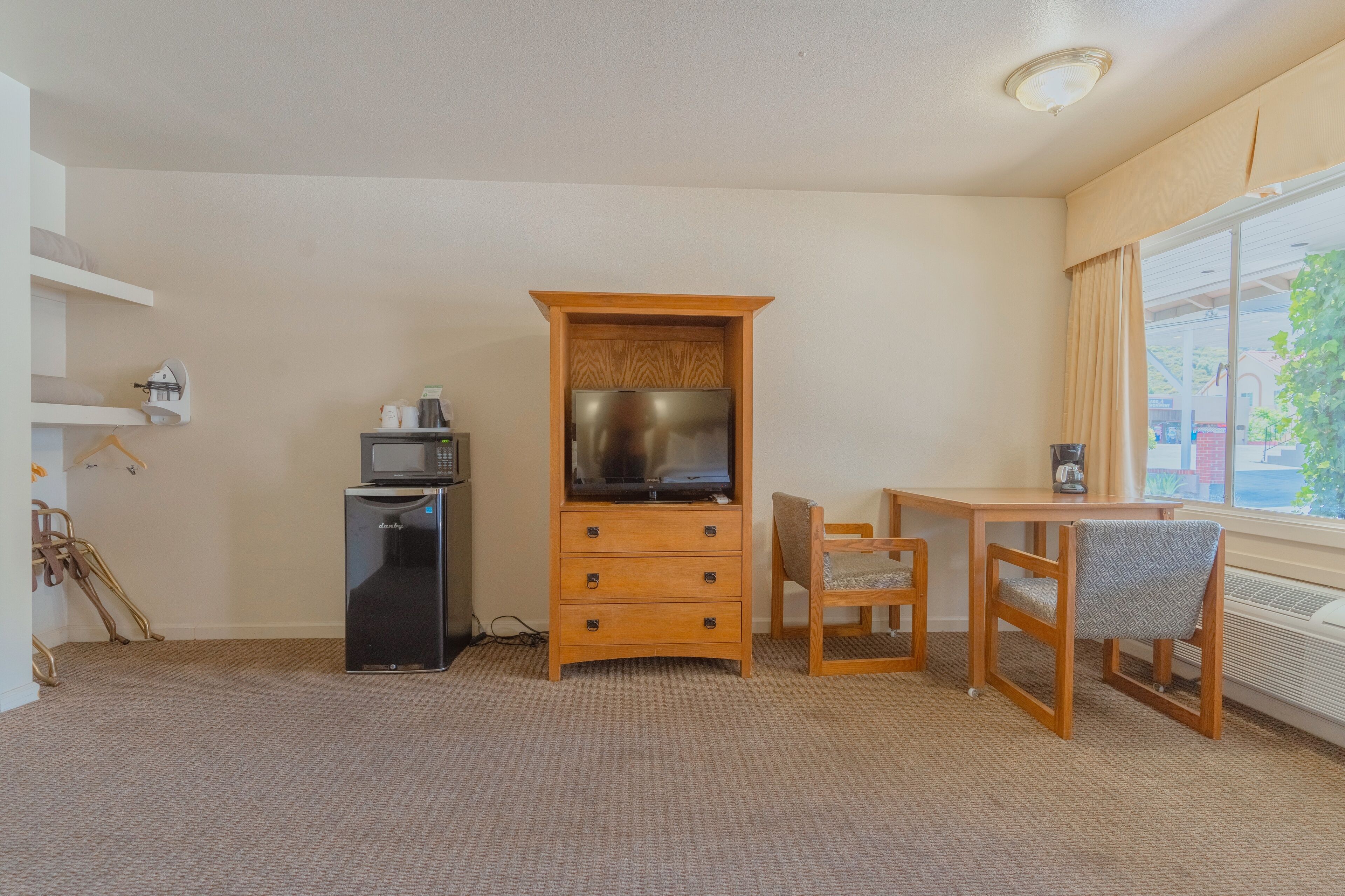 A compact room with a wooden cabinet, a small black refrigerator, a television, a wooden table with two chairs, and a window with curtains.
