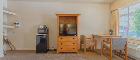 A compact room with a wooden cabinet, a small black refrigerator, a television, a wooden table with two chairs, and a window with curtains.