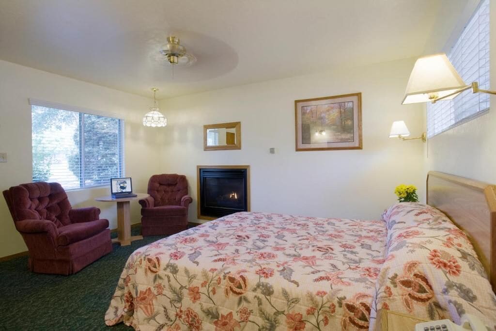 A hotel room with a bed, two armchairs, a small table, a fireplace, and a window with blinds.