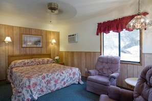 A room with a wooden paneled wall, a bed with floral bedding, a cushioned armchair, and a window with a view of snow outside.