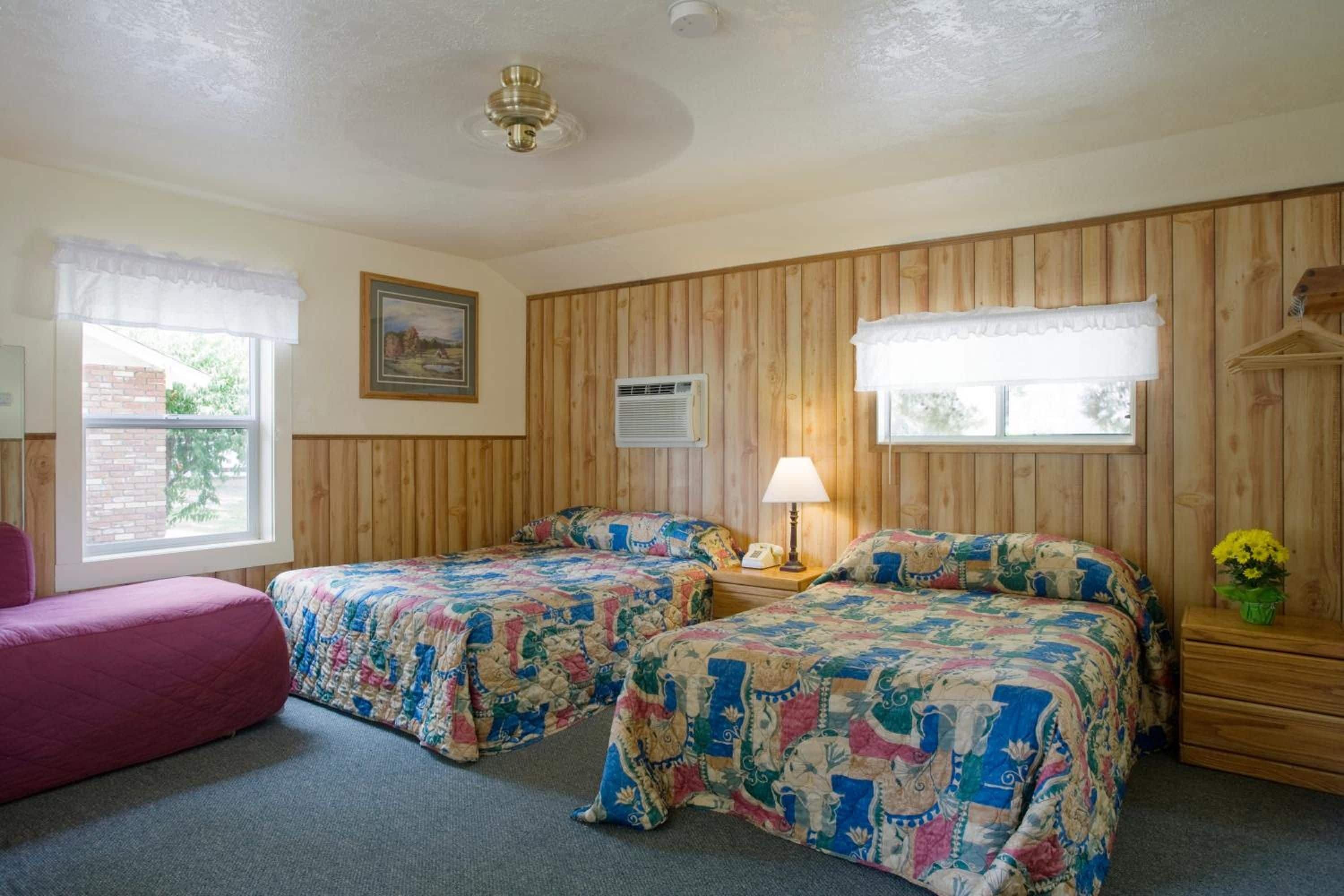 A room with two beds, a window, a painting, a lamp, and a dresser with a vase of flowers.
