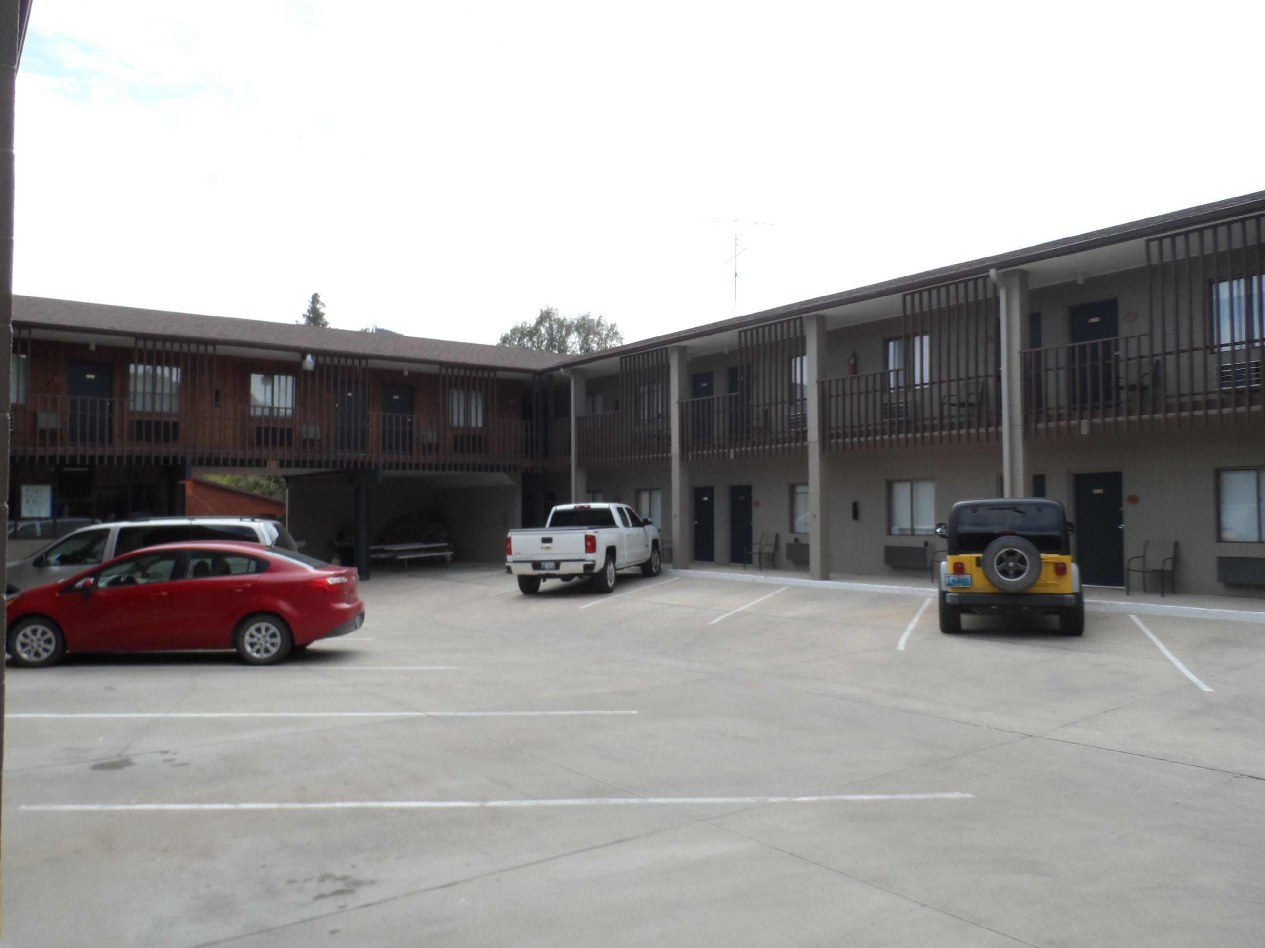 A parking lot with a building that has multiple floors and balconies. There are several vehicles parked, including a red car, a white pickup truck, and a yellow SUV.