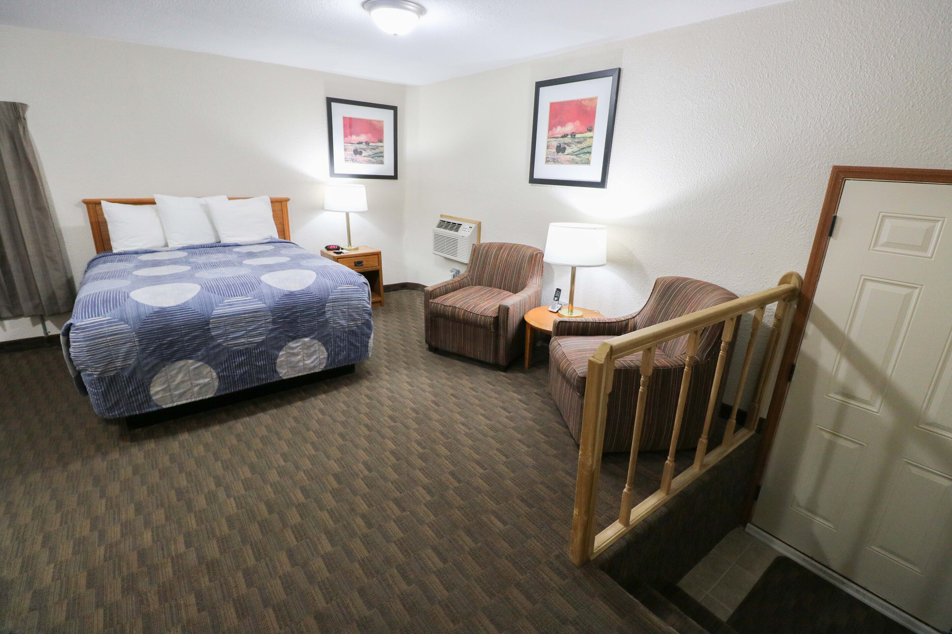A hotel room with a large bed, two armchairs, a desk with a lamp, and a door.