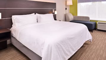 A hotel room with a large bed, a nightstand, a wall-mounted lamp, a chair, and a window with blinds.