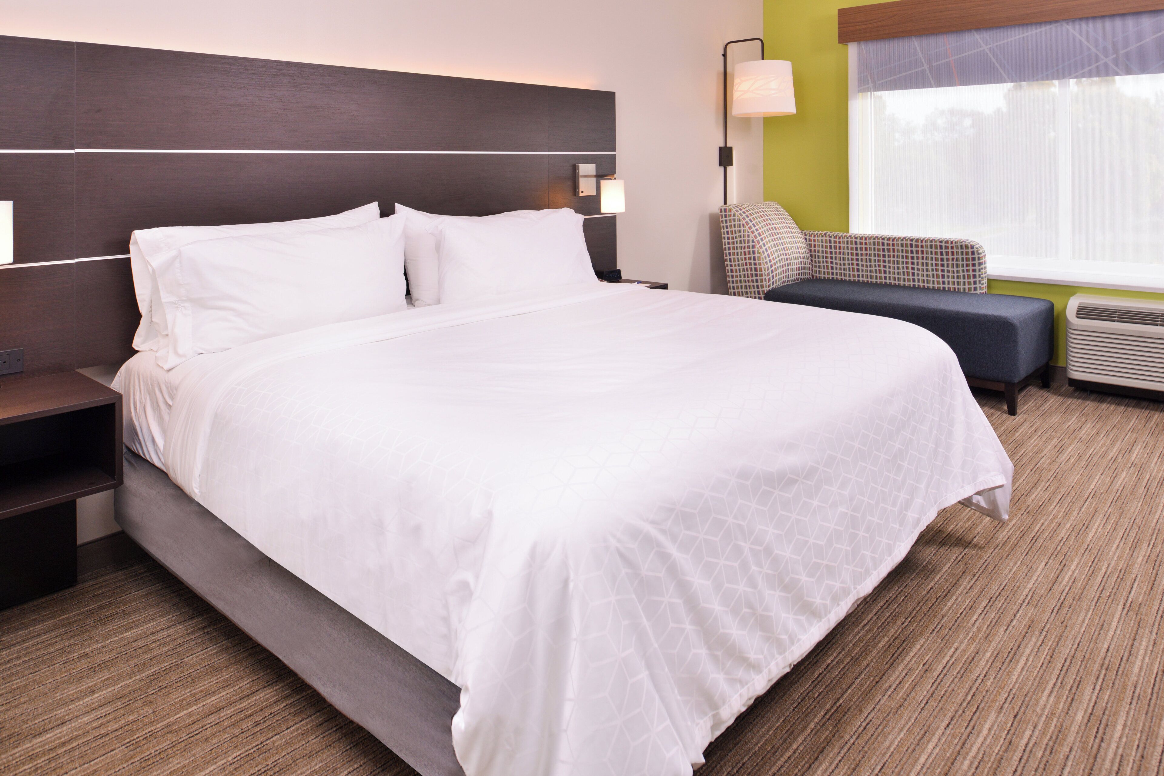 Une chambre d’hôtel avec un grand lit, une table de chevet, une chaise, une lampe et une fenêtre avec des stores.