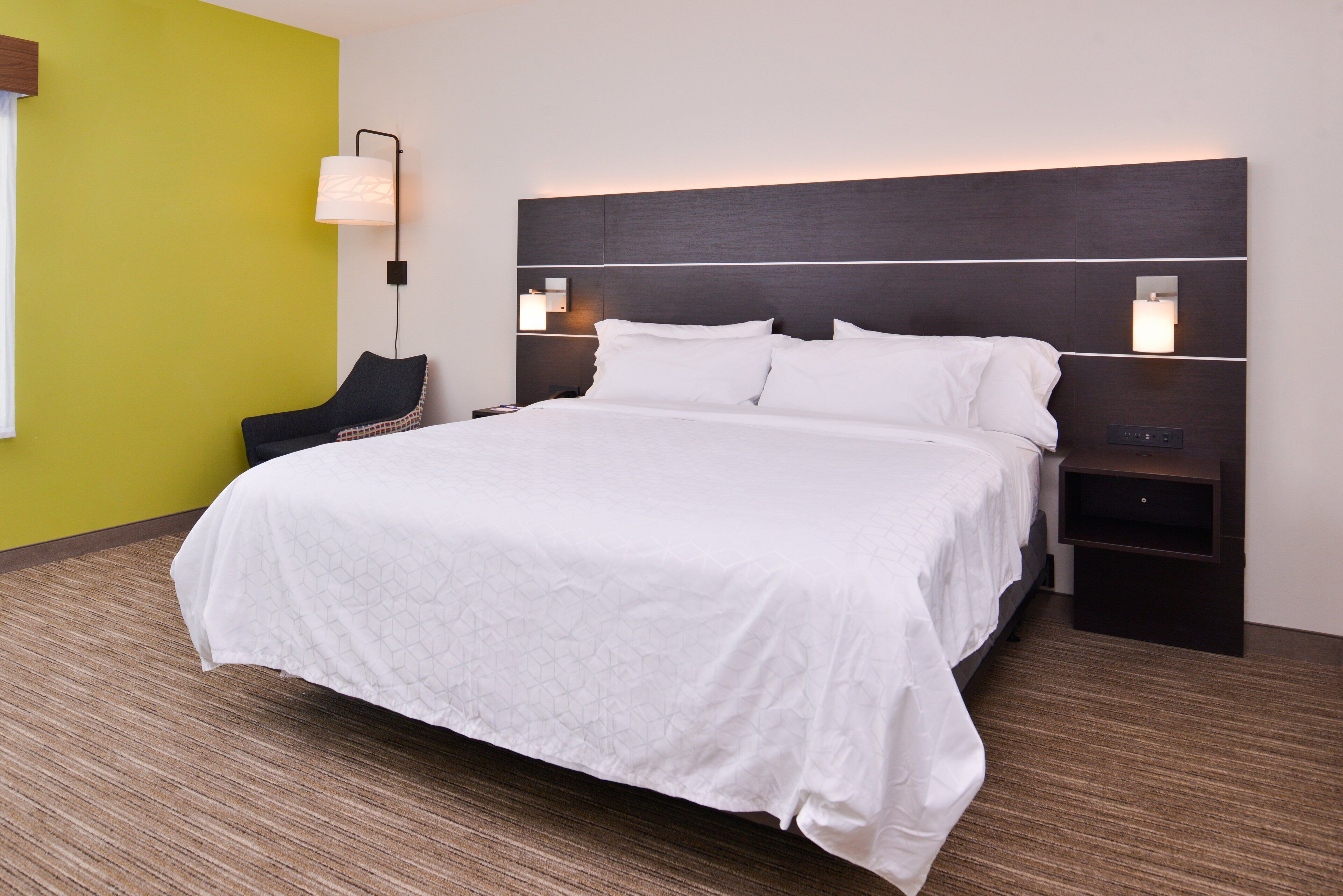 Una habitación de hotel con una cama grande, dos mesitas de noche, una silla y una lámpara de pared.