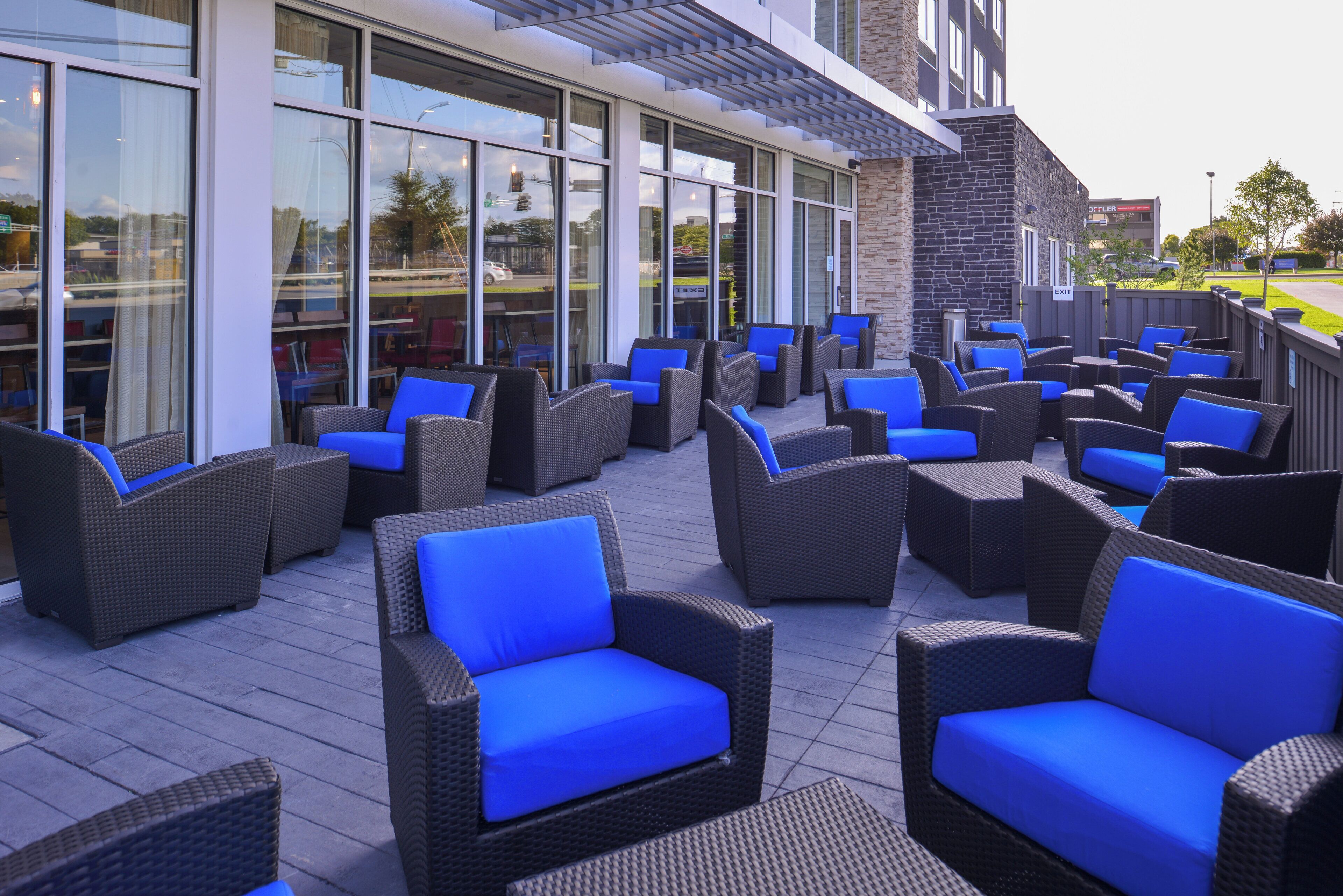 An outdoor seating area with wicker chairs and blue cushions, adjacent to a building with large windows and a stone facade.