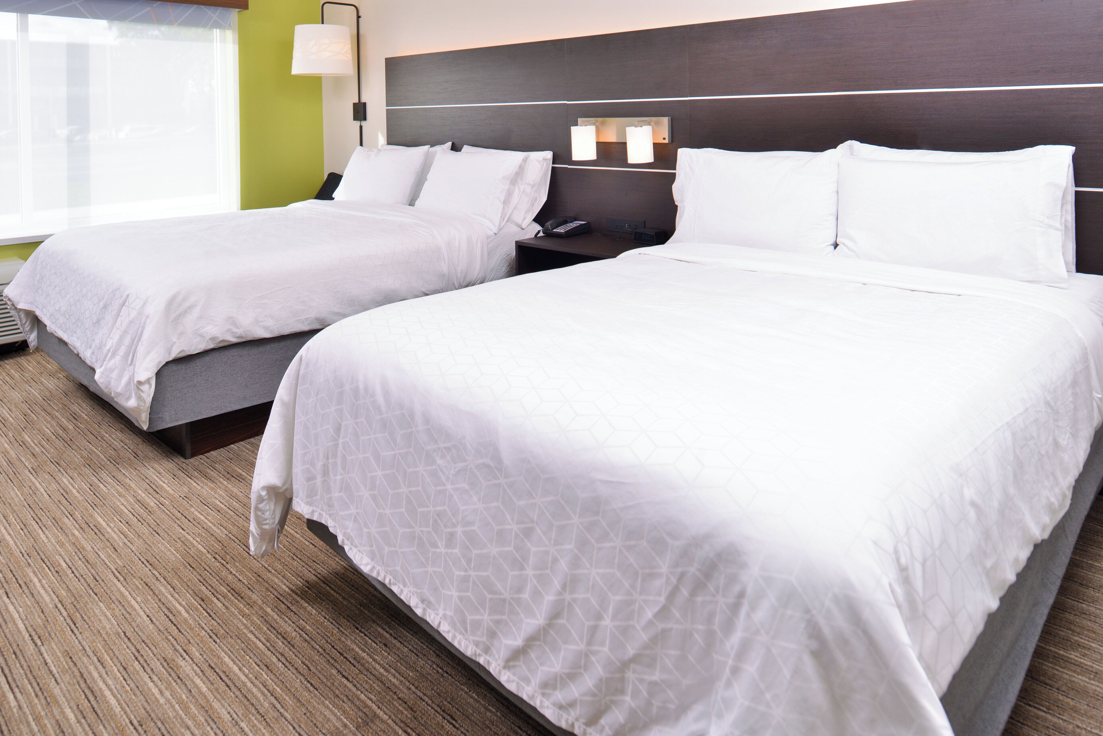 A hotel room with two beds, a nightstand, a lamp, and a window.